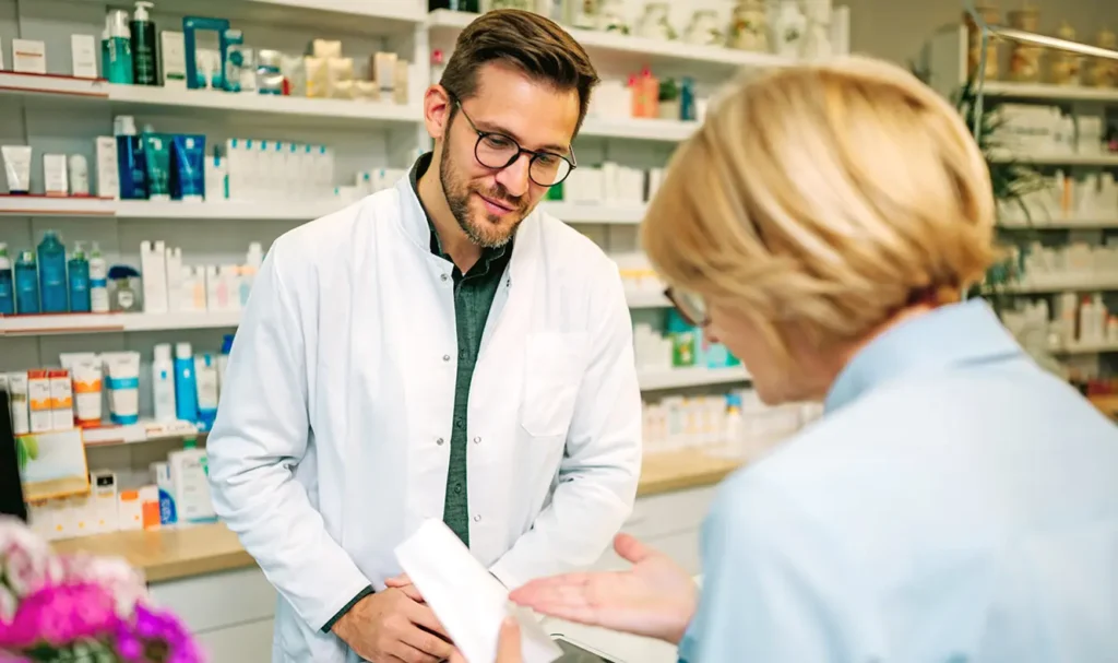 GTSN-MonthlyBlog-May Pharmacist in a white coat and glasses, smiling, assists a customer in a pharmacy. Shelves behind display various health products. Warm, professional atmosphere.